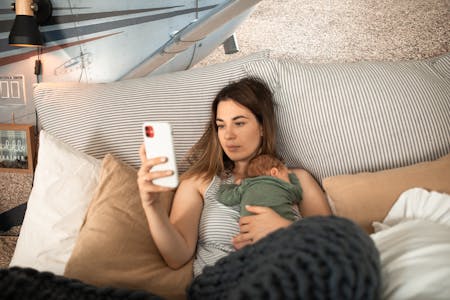 mental health statistics - woman lying in bed and holding baby while looking at her phone