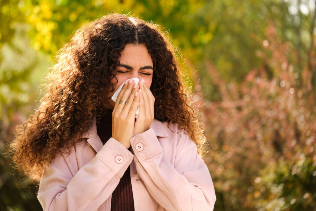 Seasonal Allergy Symptoms - Young Caucasian woman with curly hair blowing your nose in a tissue at street in Autumn.