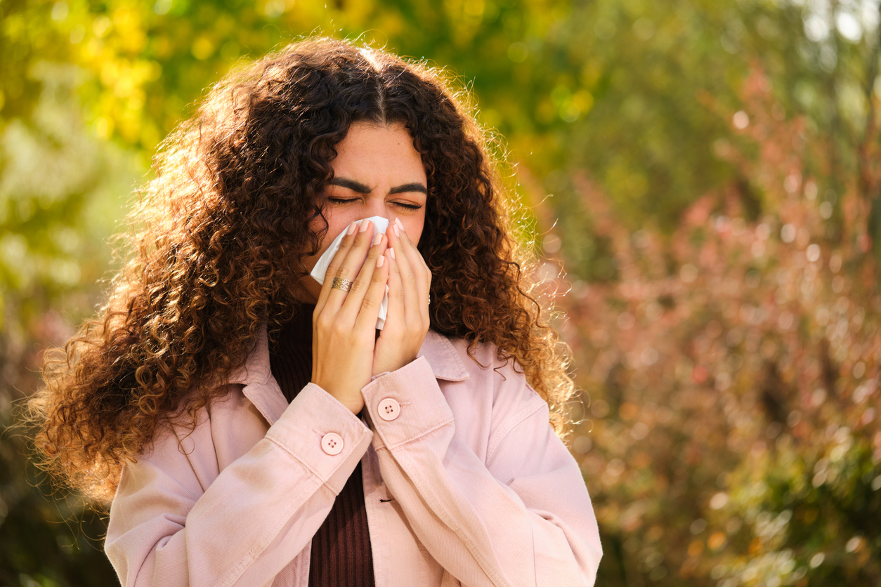 Seasonal Allergy Symptoms - Young Caucasian woman with curly hair blowing your nose in a tissue at street in Autumn.