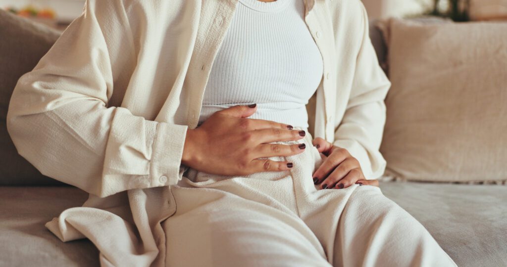 Woman, hands and stomach pain of menstruation on sofa with period cramps, bloating or pms in home. Person, abdomen and endometriosis with constipation, nausea and digestion problem on couch in lounge winter gut health woman clutching stomach